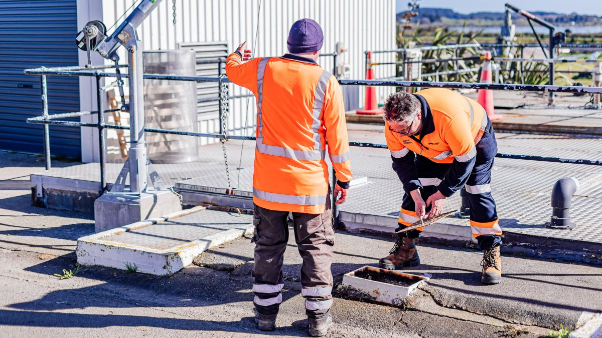 Two water workers in hi-vis inspect a drain.